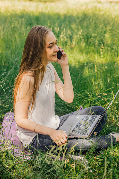 Девушка сидит на траве и работает за ноутбуком. Говорил по телефону. фрилансер. самообразование. концепция дистанционного обучения и работы на свежем воздухе - Фото, изображение