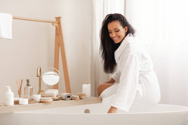 Beauty Routine. Happy Mixed Woman Sitting On Bathtub Preparing Bath Adding Sea Salt In Modern Bathroom At Home. Bodycare Cosmetics, Wellness And Spa Concept - Fotografie, Obrázek