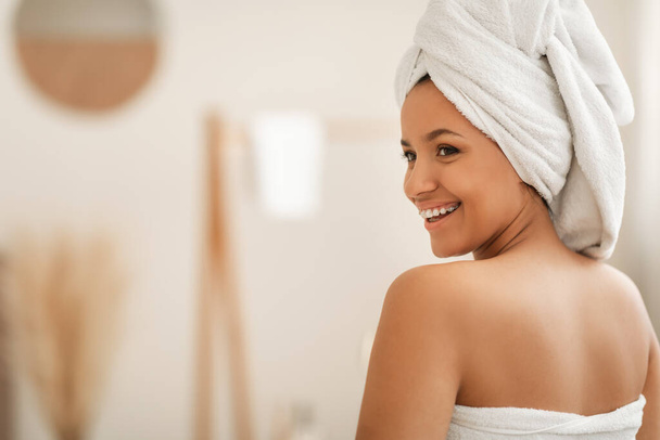 Beauty And Spa. Happy Woman Posing Wrapped In White Towel Smiling Looking Aside At Free Space Sitting Back To Camera Enjoying Bodycare Routine After Bathing In Modern Bathroom Indoors - Foto, afbeelding
