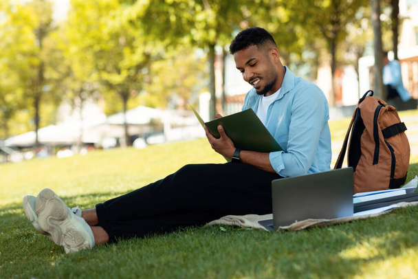 Концепція студента. Щасливий афроамериканець малює або записує нотатки, роблячи домашнє завдання, сидячи надворі в парку і посміхаючись, у вільному просторі - Фото, зображення