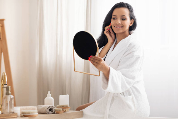 Iloinen musta nainen soveltamalla kosteusvoide katsomalla pyöreä peili nauttia kauneus rutiini ja käyttämällä kosmetiikkaa huolehtiva itsestään istuu kylpyammeessa kylpyhuoneessa sisätiloissa. Kasvojen ihonhoitotuotteet - Valokuva, kuva