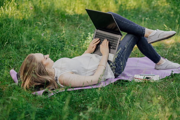On the banner, a young girl works with a laptop in the fresh air in the park, sitting on the lawn. The concept of remote work. Work as a freelancer. The girl takes courses on a laptop and smiles - 写真・画像