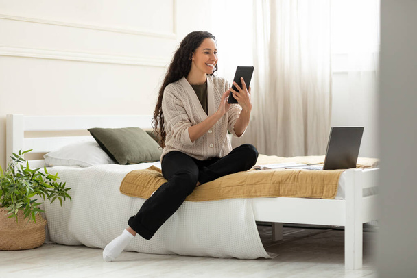 Bájos Y-generációs nő ül az ágyon, használja tabletta online munka vagy kommunikáció otthon, teljes hosszában. Pozitív fiatal nő keres internet vagy posztolás a közösségi média hordozható PC - Fotó, kép