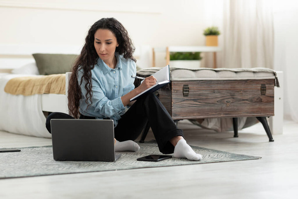 Skupiona młoda kobieta siedząca na podłodze, pracująca na laptopie, pisząca w notatniku w sypialni, wolna przestrzeń. Poważne tysiąclecia kobieta studiuje online, ogląda webinar, za pomocą komputera w domu - Zdjęcie, obraz