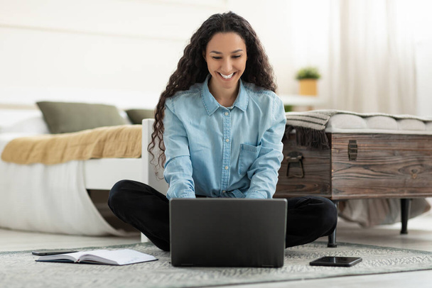 Красива молода жінка сидить на підлозі, працює на ноутбуці в спальні, повної довжини. Усміхнена тисячолітня дама використовує портативний ПК для роботи або онлайн-навчання, спілкуючись віддалено в приміщенні
 - Фото, зображення