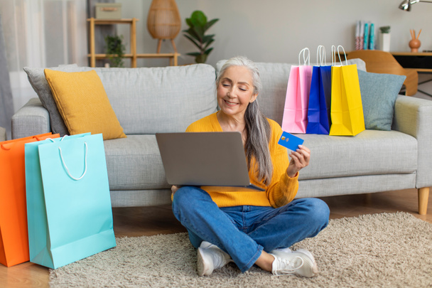 Onnellinen iloinen valkoihoinen kypsä nainen harmaat hiukset paketteja ostoksen jälkeen käyttää luottokorttia ja kannettavaa tietokonetta online-ostoksia huoneen sisustus. Eläkkeellä Shopaholic, pankki ja myynti kotona, covid-19 - Valokuva, kuva