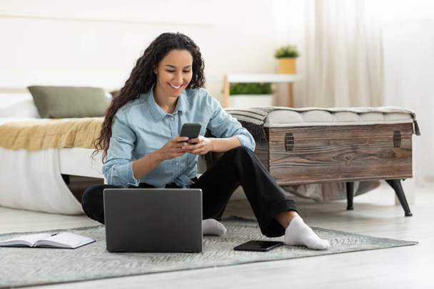 Happy young woman sitting on floor with laptop, using cell phone at home, blank space. Cheerful female with gadgets watching video or chatting online, working or studying remotely indoors - Fotografie, Obrázek