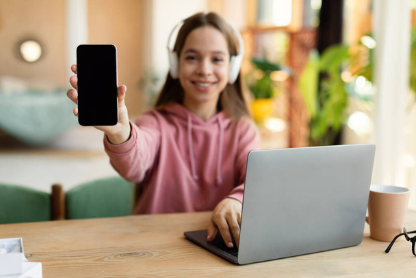 Pozytywna nastolatka demonstrująca pusty smartfon podczas nauki przy biurku z laptopem w domu, makietą, wolną przestrzenią. Kobieta student reklamy edukacyjnej aplikacji lub mobilnej stronie internetowej - Zdjęcie, obraz