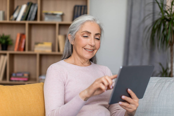 Glad smiling european senior female with gray hair have meeting remotely, chat and watch video on tablet in room interior. Work, business lesson, new normal and new app at home due covid-19 pandemic - Photo, Image