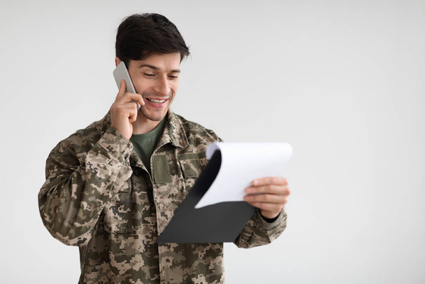 Mosolygó jóképű fiatal katona gazdaság chart és beszél a mobiltelefon, információt cserél más katonákkal, fehér stúdió háttér, panoráma fénymásolás hely - Fotó, kép