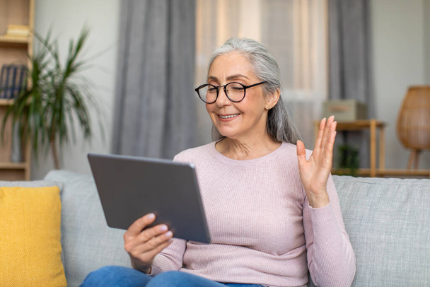 Glad european mature woman with gray hair in glasses waves her hand and looks at tablet, have meeting in living room interior. New app for video call at home, communication remotely and new normal - Photo, Image