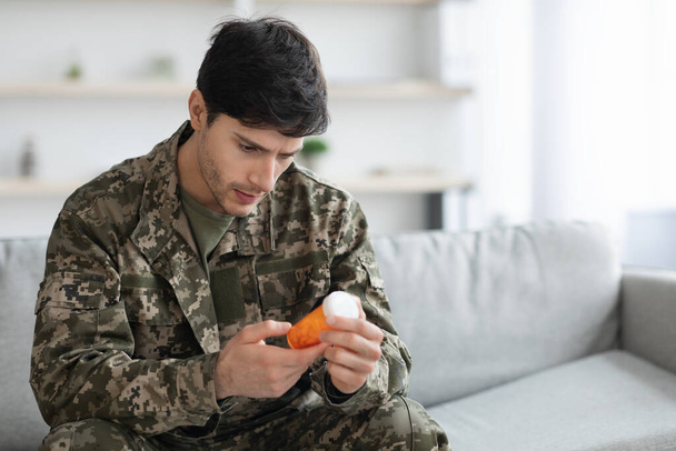 Depressed man in camouflage uniform sitting on couch, holding jar with antidepressants, suffering from depression and pain, PTSD, home interior, panorama with copy space, rehabilitation for soldiers - Photo, image