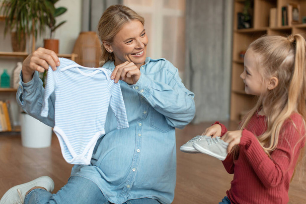 Boldog terhes nő, aki ruhákat mutat a babájának, várandós anya, idősebb lányával otthon ül a padlón, boldog család várja a babát. - Fotó, kép