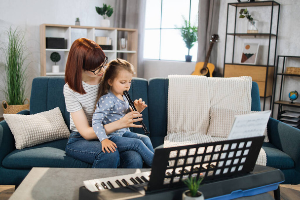 Portrait of caucasian woman and her cute little daughter learning to play flute at home. Teacher teaching pretty girl to play flute in classroom. - Foto, immagini