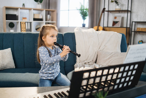 Beautiful young charming little girl smiling while playing classic flute at home. Caucasian musician girl studying to play flute indoor in light living room. - Foto, imagen