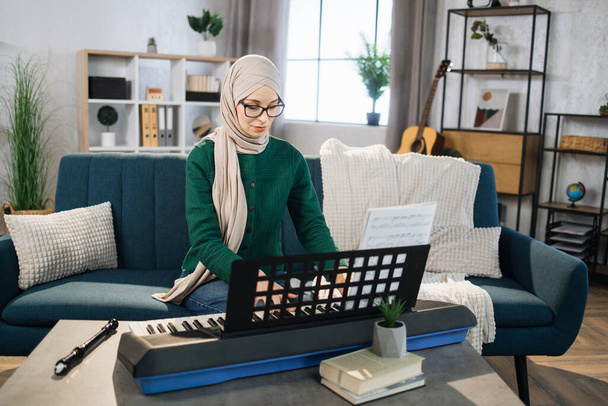 Mujer musulmana joven en hijab toca el piano en la sala de estar brillante en casa. Chica sentada en un sofá con un instrumento musical en el interior. Mujer árabe muy joven estudio para tocar el piano. - Foto, Imagen