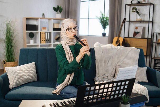 Portrait of young arab female with hijab playing on flute. Pretty muslim girl learning to play flute at home. Beautiful student woman playing musical instrument. - Fotó, kép