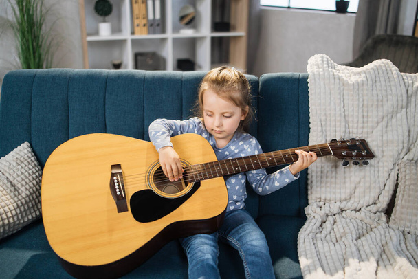 Beautiful young charming little girl smiling while playing classic guitar at home. Caucasian musician girl studying to play guitar indoor in light living room. - 写真・画像