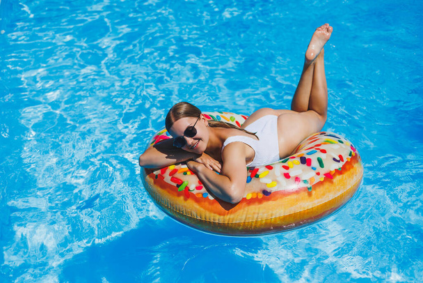 Riposa in piscina. Giovane donna felice in costume da bagno, occhiali da sole e anello di gomma gonfiabile galleggiante in acqua blu. Vacanze estive di lusso nella piscina del resort termale - Foto, immagini