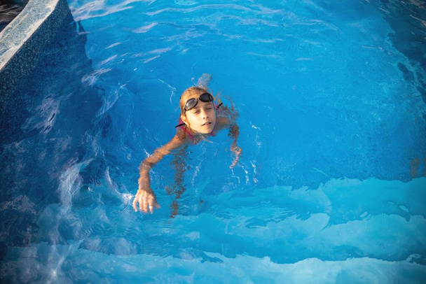 A little girl in a bright pink swimsuit with transparent black goggles for swimming, straightens her goggles and then dives into a deep blue pool with a chimta of clear water - Photo, image