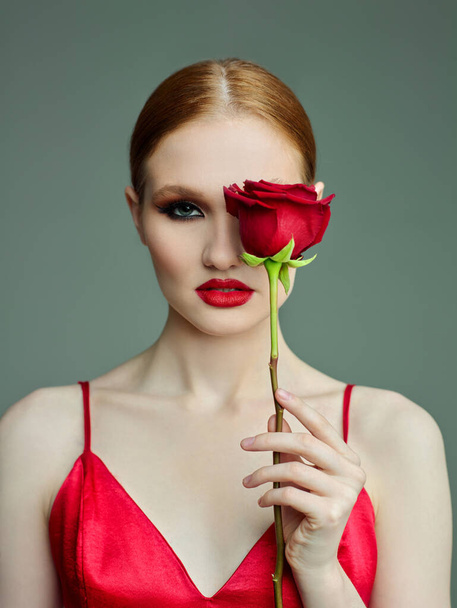 Yüzünün yanında kırmızı dudakları ve büyük kırmızı bir gül olan genç bir kadının portresi. Kırmızı elbiseli mankenle güzel bir poz.. - Fotoğraf, Görsel