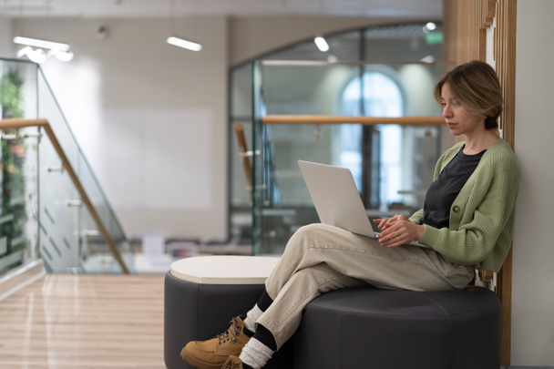 Focused pensive middle-aged woman sitting on ottoman with laptop, scandinavian lady freelance writer working remotely in coworking space, serious concentrated mature female remote worker in library - Foto, imagen