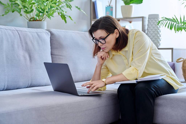 Mature businesswoman working on couch, using laptop making notes in business notebook. Confident middle aged female working from home office remotely. Business job freelancing, technology, 40s people - Photo, image