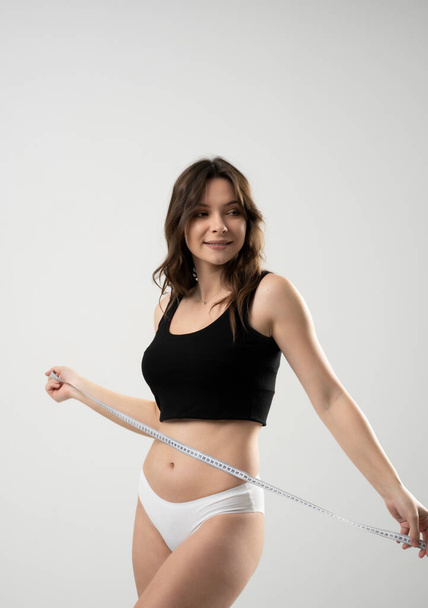 Cheerful beautiful sexy brunette woman girl in black top and white shorts with a measuring tape on a white background - Φωτογραφία, εικόνα
