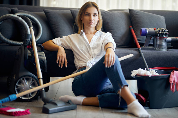 Mujer sentada en el suelo cerca del sofá con muchas herramientas de limpieza - Foto, Imagen