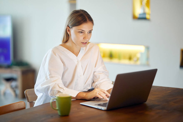 白いシャツの美しい女性はテーブルでコーヒーを飲みながらノートパソコンで働いています - 写真・画像