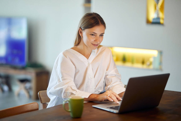 Красива жінка в білій сорочці працює з ноутбуком вдома з чашкою кави за столом
 - Фото, зображення