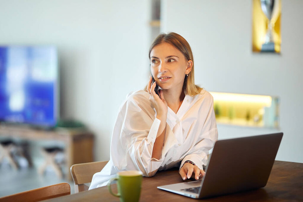 Hermosa mujer con camisa blanca está teniendo una llamada telefónica en casa con un ordenador portátil y una taza de café en la mesa. - Foto, imagen