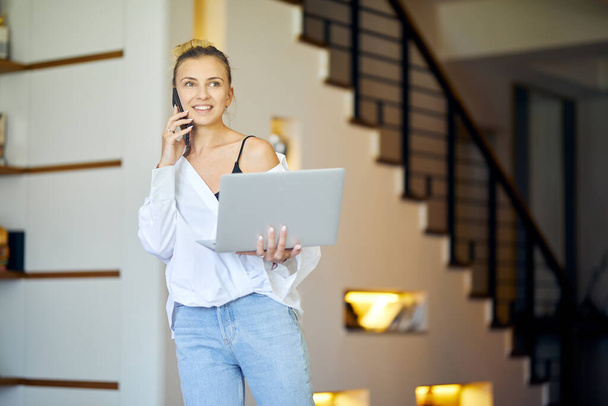 Una mujer en casa con un portátil en la mano está teniendo una llamada telefónica y oliendo. Escaleras en el fondo. De interior - Foto, imagen
