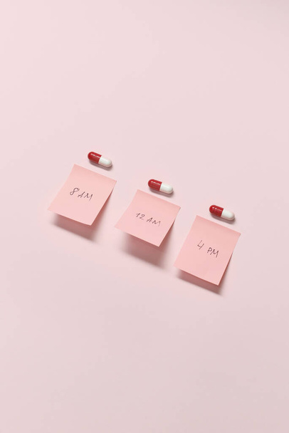 Red and white pharmaceutical capsules and sticky notes with time of medication over pink background, copy space. - Fotografie, Obrázek