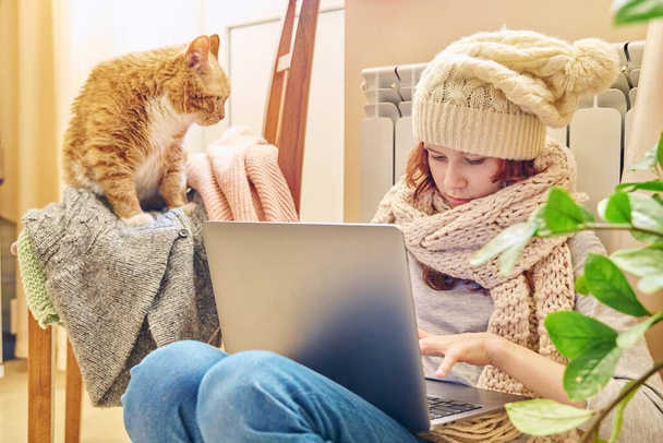Preteen girl in knitted scarf hat is warming near heating radiator using laptop for leisure study with ginger cat lying on chair. Cold autumn winter season, home lifestyle, kids concept - Φωτογραφία, εικόνα