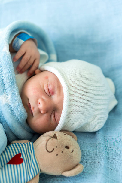 Close-up of Newborn Baby Face Portrait Early Days Sleeping With Tady Bear On Blue Background. Child At Start Minutes Of Life on Hat. Infants, Childbirth, First Moments Of Borning, Beginning Concept - 写真・画像