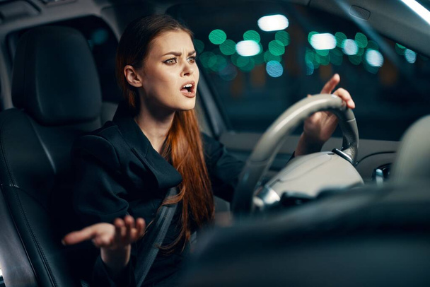 egy vízszintes fotó egy nőről, aki egy autó volánjánál ül, és a szája szélesre tárva, meglepetten tárja szét a karját az oldalán. Fényképezés éjszaka. Kiváló minőségű fénykép - Fotó, kép