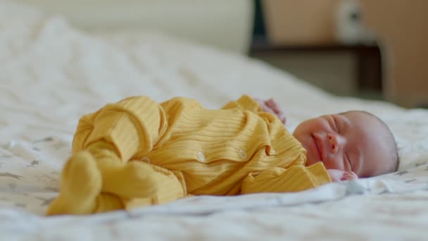 4k Filmmaterial Winziges Neugeborenes in gelbem Overall schläft auf dem Rücken mit erhobenen Armen und lächelt im Schlaf. Kleines süßes Baby liegt auf einem großen Elternbett oder wacht mit glücklichem Lächeln auf  - Filmmaterial, Video