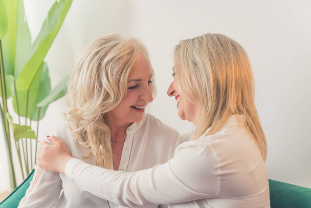 Primer plano de interior de dos mujeres caucásicas de mediana edad con el pelo rubio y camisas blancas sentadas en un sofá abrazándose y riendo. Foto de alta calidad - Foto, imagen