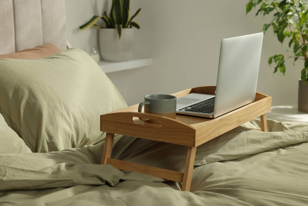 Wooden tray with modern laptop and cup of aromatic drink on bed indoors - Photo, Image