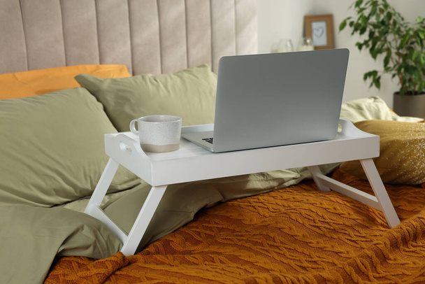 White tray with modern laptop and cup of aromatic drink on bed indoors - Foto, Imagen