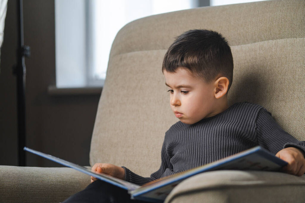 Bambino che legge un libro seduto sul divano, a casa. Istruzione e sviluppo precoce dei bambini. - Foto, immagini