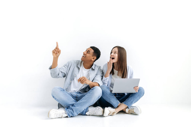 Glückliches multiethnisches Paar, ein hispanischer Mann und eine kaukasische Frau, mit Laptop, schauen zur Seite, der Kerl zeigt mit der Hand auf etwas, während er auf dem Boden auf weißem, isoliertem Hintergrund sitzt, lächelt - Foto, Bild