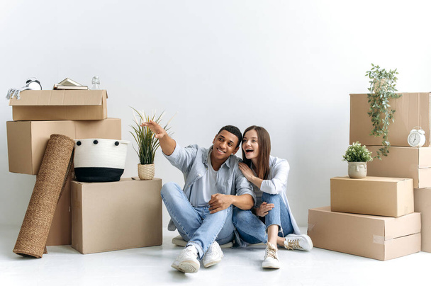 Счастливая молодая многорасовая пара, кавказская женщина и латиноамериканец, сидят на полу между коробками с вещами в своем новом доме или арендованной квартире, планируя свой дом будущего дизайна, улыбаются - Фото, изображение