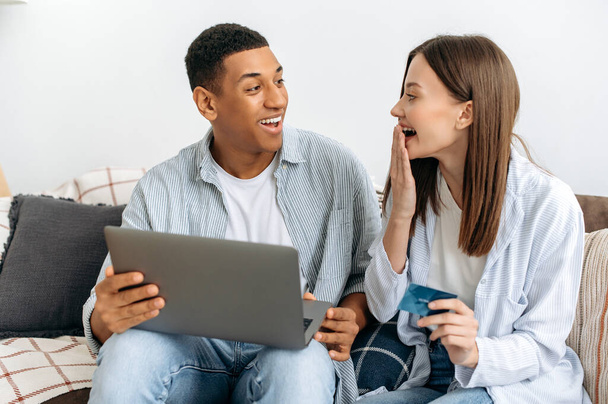 Aufgeregt glücklich stilvolle multirassische Paar in Liebe, Ehegatten, sitzen auf dem Sofa in einem Wohnzimmer, mit Laptop und Kreditkarte für Online-Shopping, Lieferung, schauen einander erstaunt, lächelnd - Foto, Bild