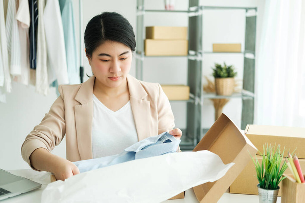 Jeune femme d'affaires asiatique startup entrepreneur propriétaire de PME ramasser une chemise jaune avant de l'emballer dans une boîte intérieure avec un client. Idées d'affaires en ligne et freelance. - Photo, image