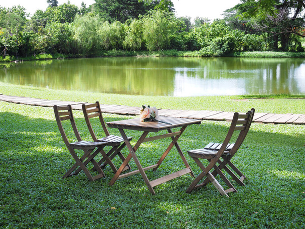 Tabel de madera y sillas colocadas en el patio de hierba verde cerca del lago en el parque en el día soleado. - Foto, Imagen