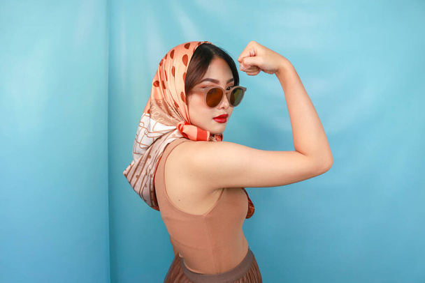 Retro concept of a young Asian strong woman showing her biceps isolated by a blue background - Photo, Image