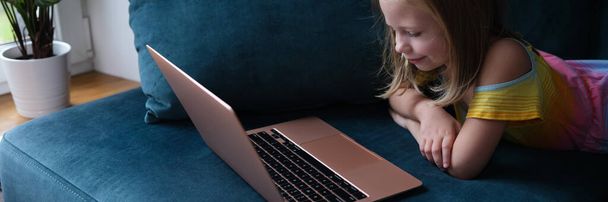 Küçük bir kız dizüstü bilgisayarla kanepede yatıyor, yakın plan. Çocuk bir video konferansına katılıyor, ebeveynlerle uzaktan iletişim kuruyor. - Fotoğraf, Görsel