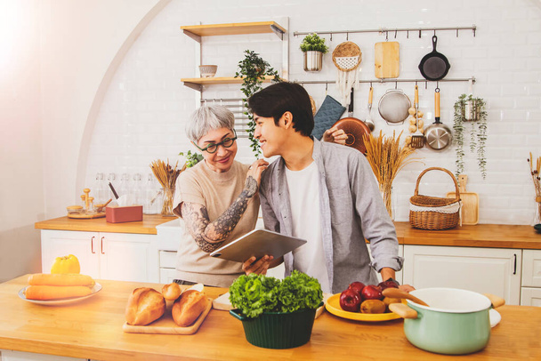 Couple between an old woman tattooed on the arm and handsome childhood boy who bond and love each other well, embrace close care, cook in the kitchen with joy : Age difference and love concept. - Foto, afbeelding